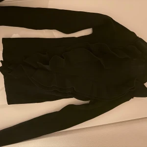 Svart ribbad polotröja med volang - Snygg svart polotröja med ribbad struktur och dekorativa volanger framtill. Tröjan är tjock i materialet vilket både håller värme och det ser lyxigare ut. Aldrig använd.
