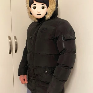Svart pufferjacka med päls från SikSilk - Svart pufferjacka från SikSilk med fluffig päls runt huvan och snygga detaljer som knappar och dragkedja på ärmen. Jackan har lång ärm, flera fickor och en stilren logga på bröstet. Perfekt för kalla dagar och riktigt skön att bära.