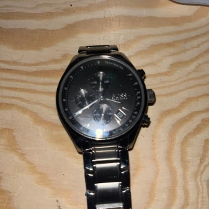 Silverfärgad kronografklocka från BOSS - Stilren och robust kronografklocka från BOSS i silverfärgat rostfritt stål. Klockan har svart urtavla med flera små ur och datumvisning, samt länkarmband med BOSS-logga på spännet. Perfekt accessoar för dig som gillar klassisk och modern design. OBS: Nytt batteri får du köpa själv 