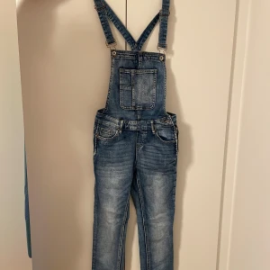 Blå jeansbyxdress med hängslen - Säljer en snygg blå jeansbyxdress med klassiska hängslen och fickor både fram och bak. Byxdressen har en cool tvättad look och justerbara axelband. Perfekt för dig som gillar avslappnad stil och vill sticka ut lite extra. Oanvänd 