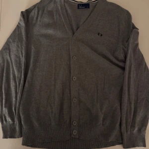 Grå kofta från Fred Perry - Snygg grå kofta från Fred Perry med v-ringad hals och knappar hela vägen framtill. Diskret logga broderad på bröstet och ribbade muddar vid ärmslut och nederkant. Perfekt för lager-på-lager och chill stil. Pris går diskutera vid snabb affär, använd sparsamt 