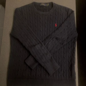 Mörkblå kabelstickad tröja Polo Ralph Lauren - Kabelstickad mörkblå tröja från Polo Ralph Lauren med klassisk rund halsringning och röd broderad logga på bröstet. Tröjan har långa ärmar och ribbade muddar, perfekt för en stilren och chill look.