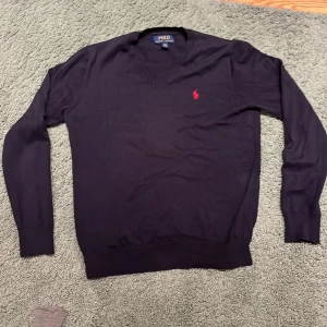 Svart v-ringad tröja Polo Ralph Lauren - Svart v-ringad tröja från Polo Ralph Lauren med rött broderat logomärke på bröstet. Tröjan har långärmad design och är tillverkad i mjuk bomull. Ribbstickade muddar vid ärmslut och nederkant ger en clean och klassisk look.