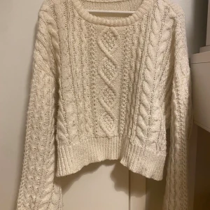 Krämvit  stickad tröja - Säljer en krämvit stickad  tröja med snyggt flätmönste och ribbade muddar. Tröjan har lång ärm och rund halsringning, perfekt för dig som gillar mysiga och trendiga plagg med lite oversized känsla.