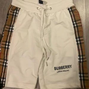  Burberry shorts - Snygga vita shorts från Burberry med klassiska rutiga paneler längs sidorna och logga framtill. De har bekväm resårmidja med snörning och är perfekta för en trendig och avslappnad stil.