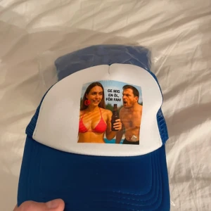 Roliga - Säljer en blå och vit truckerkeps med mesh-bak och justerbar snapback. Framsidan har ett färgglatt tryck med texten 'GE MIG EN ÖL, FÖR FAN!' och motiv av två personer vid en pool. Perfekt för sommarens alla sköna dagar.