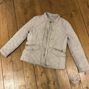 Beige quiltad jacka med dragkedja - Snygg beige quiltad jacka från Zara med ståkrage och långa ärmar. Jackan har två fickor med dragkedja och knappdetaljer framtill. Perfekt för dig som gillar stilren och klassisk look.
