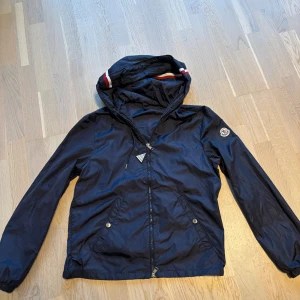 Mörkblå vindjacka från Moncler - Snygg mörkblå vindjacka från Moncler med huva och dragkedja. Jackan har Moncler-logga på ärmen, två fickor med tryckknappar och coola rödvita detaljer på huvan. Tillverkad i lätt och glansig polyester, perfekt för blåsiga dagar.