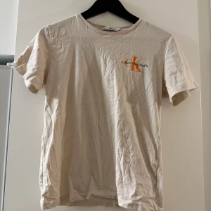 Beige t-shirt från Calvin Klein Jeans - Snygg beige t-shirt från Calvin Klein Jeans med orange CK-logga tryckt på bröstet. Klassisk rund hals och korta ärmar. Tillverkad i mjuk bomull som känns skön mot huden. Perfekt för dig som gillar stilrena och enkla plagg med en touch av branding.