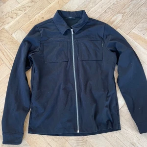 Svart overshirt från Jack & Jones M - Stilren svart overshirt från Jack & Jones Premium med raka linjer, två bröstfickor och hel dragkedja framtill. Jackan har klassisk krage och är tillverkad i ett mjukt bomullsmaterial. Perfekt för lager-på-lager och enkel att matcha.