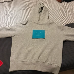 Grå hoodie Acne Studios med patch - Grå hoodie från Acne Studios med huva och utbytbar broderi-patch på bröstet i blått, samt extra röd patch. Tröjan har normal passform och är tillverkad i mjuk bomull. Passar både M och S skulle jag säga!