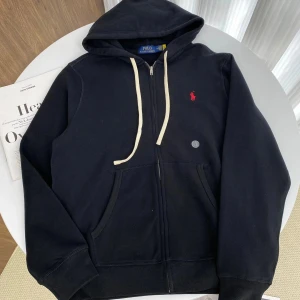 Svart hoodie från Polo Ralph Lauren - Svart hoodie från Polo Ralph Lauren med vit snörning och dragkedja framtill. Tröjan har huva, två fickor och den klassiska röda Polo-loggan broderad på bröstet. Tillverkad i mjuk bomull, perfekt för chill dagar och streetwear.