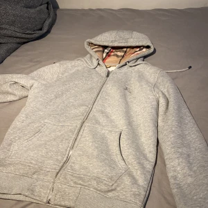 Grå hoodie från Burberry med dragkedja - Snygg grå hoodie från Burberry med klassiskt rutigt foder i huvan och insidan. Tröjan har dragkedja, broderad logga på bröstet och två fickor framtill. Perfekt för dig som gillar stilrena och exklusiva plagg med en twist. 