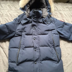 Mörkblå dunjacka från Canada Goose - Säljer en mörkblå dunjacka från Canada Goose med pälskantad huva och klassisk logga på ärmen. Jackan har långa ärmar, stora fickor med dragkedja och knappar framtill. Perfekt för kalla vinterdagar och har en riktigt schysst, puffig look.