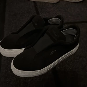 Svarta sneakers från Axel Arigato - Svarta slip-on sneakers från Axel Arigato med vit platt sula och minimalistisk design. Skorna har ett inlägg där bak som är för att motverka skavsår få dem glappade en del på mig och vilket är därför jag säljer. Använda ca 2 gånger💓orginalpris ca 2500 (osäker på om inläggen går att ta bort)+finns låda och dustbag