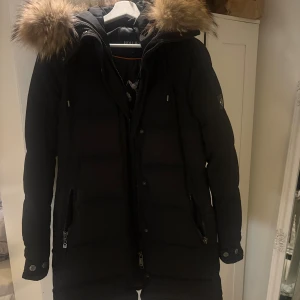 Svart pufferjacka Hollies med päls - Svart pufferjacka från Hollies med stor fluffig päls på huvan och snygga detaljer som dragkedjor och knappar. Jackan har lång ärm, emblem på armen och coolt Arctic Expedition-tryck på insidan. Perfekt för kalla vinterdagar och riktigt stilren look.