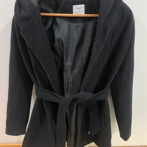 Svart kappa med huva från Vero Moda - Svart kappa från Vero Moda med huva och bälte i midjan. Jättebra skick