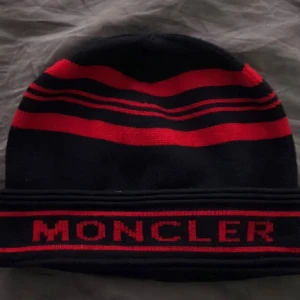 Svart och röd Moncler mössa - Snygg svart mössa från Moncler med röda ränder och tydlig logga framtill. Mössan har en klassisk form och är stickad i mjukt material som håller dig varm. Perfekt för dig som vill ha en cool accessoar med streetkänsla.