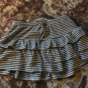 Randig volangkjol från Gina Tricot - Superfin kort kjol från Gina Tricot med svartvita ränder och volangdetaljer. Kjolen har en söt knytning i midjan och är gjord i ett lätt, skönt material som passar perfekt till varma dagar. Den har en avslappnad och trendig vibe.