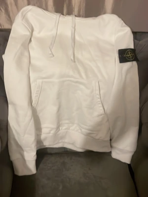 Vit hoodie från Stone Island - Säljer en clean vit hoodie från Stone Island med klassisk patch på ärmen. Hoodien har huva med dragsko och en stor magficka. Tillverkad i mjukt bomullsmaterial och har ribbade muddar vid ärmslut och nederkant. Perfekt för dig som gillar streetwear och snygga detaljer.