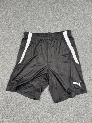 Svarta träningsshorts från Puma - Snygga svarta träningsshorts från Puma med vita detaljer på sidorna och klassisk Puma-logga på vänster ben. Shortsen har elastisk midja och är tillverkade i ett lätt och svalt polyestermaterial, perfekta för sport och aktivitet.