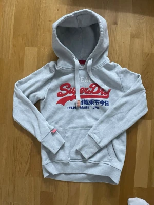 Grå Superdry hoodie med tryck - Snygg grå hoodie från Superdry med stor röd och blå logga på bröstet och japansk text. Klassisk känguruficka framtill, ribbade muddar och dragsko i huvan. Mjuk bomullsblandning som känns skön mot huden. Perfekt för en avslappnad streetwear-look.