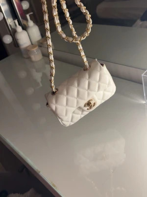 Vit quiltad Chanel handväska - Supersnygg vit quiltad handväska från Chanel med klassiskt guldigt CC-spänne framtill. Väskan har en kedjerem med insytt vitt läder och den ikoniska rutmönstrade designen. Perfekt för dig som vill ha en lyxig och tidlös accessoar till din outfit.