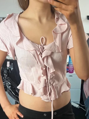 Ljusrosa volangblus med knyt - Supersöt ljusrosa topp med volangdetaljer och knyt framtill. Kortärmad och croppad modell med djup ringning och rynkad midja. Perfekt för en romantisk look och till sommaren. Materialet känns lätt och luftigt.