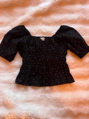 Svart prickig topp från H&M - Söt svart topp från H&M med vita prickar och puffiga korta ärmar. Toppen har smock i midjan och vid halsen för en snygg passform. Perfekt med typ kjol eller jeans. I storlek 158💕