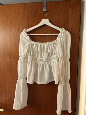 Vit offshoulder blus med volanger - Superfin vit offshoulder blus med långa, vida ärmar och smockdetaljer vid byst och midja. Blusen har en luftig och flowy passform med volanger och resår vid halsringningen. Perfekt för en trendig och avslappnad look.