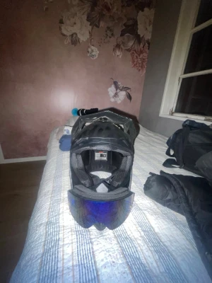 Svart hjälm med visir och goggles - Säljer en svart hjälm med skärm och blåtonade goggles. Hjälmen har ett sportigt utseende med ventilationshål och justerbar skärm. Gogglesen har ett vitt och svart mönstrat band. Perfekt för dig som gillar fartfyllda aktiviteter.