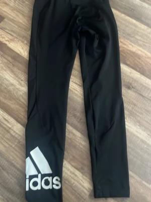 Svarta leggings från Adidas - Snygga svarta leggings från Adidas med stort vitt logotryck på ena benet. De har en tight passform och är tillverkade i stretchigt material som passar perfekt till träning eller chill. Klassisk midja och långa ben. Fast det på Adidas har lite gått bort men inget man tänker på.