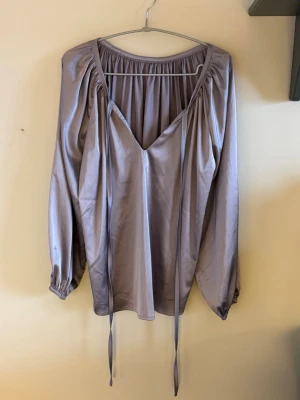 Ljuslila satintopp med v-ringning - Snygg ljuslila blus i satinliknande material med djup v-ringning och långa knytband framtill. Blusen har puffiga långärmar med resår vid ärmslut och en avslappnad passform. Perfekt för dig som gillar trendiga och feminina plagg.