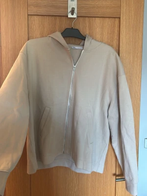Beige ziphoodie från Zara - Stilren beige hoodie med dragkedja från Zara. Modellen har huva, två fickor framtill och är tillverkad i mjukt bomullstyg. Perfekt för en avslappnad look och enkel att matcha med andra plagg.