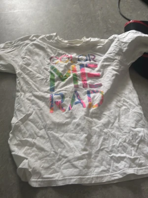 Vit t-shirt med färgglatt tryck - Vit t-shirt med texten 'COLOR ME RAD' i stora bokstäver med färgglada detaljer. Klassisk passform och rund halsringning. Perfekt för dig som gillar färg och vill sticka ut lite extra.