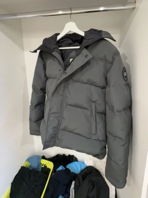 Mörkgrå dunjacka från Canada Goose - Snygg mörkgrå dunjacka från Canada Goose med avtagbar huva och diskret logga på ärmen. Jackan har dragkedja och framtill samt två stora fickor. Perfekt för kalla vinterdagar tack vare det tjocka, vadderade materialet.