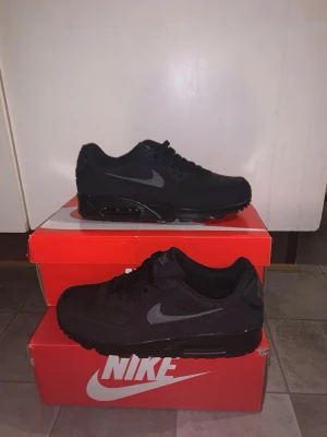 Nike Air Max 90 helsvarta sneakers - Snygga helsvarta Nike Air Max 90 sneakers med klassisk siluett. Skorna har en ovandel i mesh och syntet, svart sula med synlig Air-enhet och mörkgrå Swoosh på sidan. Perfekta för dig som gillar stilrena och tidlösa sneakers.