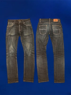 Nudie Selvage Grim Tim  - Säljer ett par mörkblå jeans från Nudie Jeans med klassisk femficksdesign och kontrastsömmar. Jeansen har raka ben och tydliga slitningar på framsidan och baksidan för en cool, använd look. Perfekta för dig som gillar streetstyle.