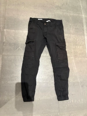 Svarta cargobyxor med fickor - Snygga svarta cargobyxor med flera stora fickor på sidorna och resår vid bensluten. Byxorna har en avslappnad passform och klassisk cargostil, perfekta för en streetwear-look. Materialet ser ut att vara bomull eller bomullsblandning.