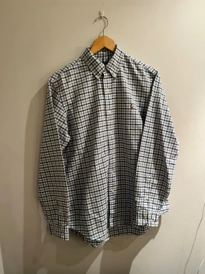 Rutig skjorta från Ralph Lauren - Säljer en klassisk rutig skjorta från Ralph Lauren i blått och vitt. Skjortan har lång ärm, knappar hela vägen och en liten broderad logga på bröstet. Tillverkad i mjuk bomull och har en normal passform. Perfekt för dig som gillar stilrena och tidlösa plagg.