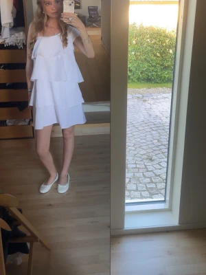 Vit volangklänning - Söt vit kortklänning med volanger och axelbandslös design. Perfekt för varma dagar och somriga tillfällen. Klänningen har en luftig passform och är tillverkad i ett lätt material. Från Gina Young storlek XS. 