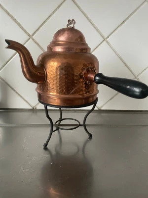 Antik Koppartekanna med svart handtag - Säljer en klassisk tekanna i hamrad koppar med svart handtag och böjd pip. Kannan står på ett dekorativt stativ i svart metall och har ett lock med liten knopp. Perfekt som inredningsdetalj eller för servering av te.