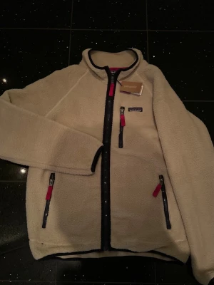 Beige teddyjacka från Patagonia - Mysig beige teddyjacka från Patagonia med svarta detaljer och röda dragkedjor. Jackan har hel dragkedja framtill, två sidofickor och en bröstficka. Tillverkad i fluffigt teddy-material som håller dig varm och bekväm. Perfekt för kyliga dagar.