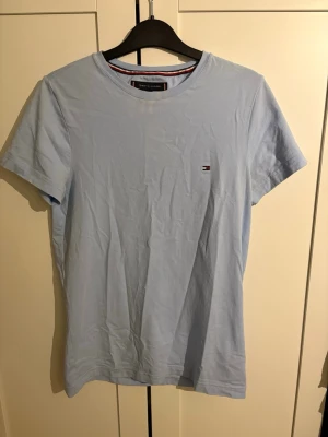 Ljusblå t-shirt från Tommy Hilfiger - Snygg ljusblå t-shirt från Tommy Hilfiger i slim fit-modell. Tillverkad i ekologisk bomull och har en liten broderad logga på bröstet. Perfekt för en stilren och enkel look. OBS! De är gul / vit fläckig vid kragen men syns ej lika tydligt i person som på bild