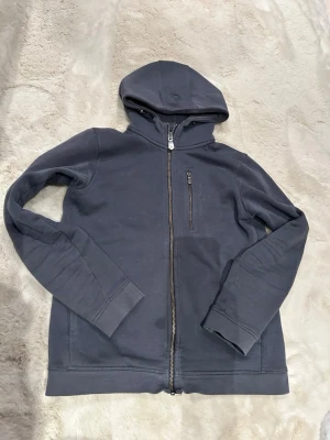 Mörkgrå hoodie från Sail Racing - Mörkgrå hoodie med dragkedja från Sail Racing. Tröjan har en huva, bröstficka med dragkedja och broderad logga på bröstet. Tillverkad i mjukt material och har ribbade muddar vid ärmslut och nederkant. Perfekt för en avslappnad och sportig stil.