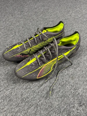 Puma ultra 5 fotbollsskor  - Säljer ett par snygga fotbollsskor från Puma i svart med neon-gula detaljer och tunna rosa inslag. Skorna har snörning, låg profil och är tillverkade i ett lätt syntetmaterial för bästa känsla på planen. Perfekta för dig som vill sticka ut på matchen!