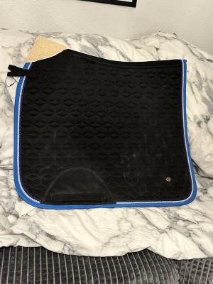 Svart schabrak med blå kant - Säljer ett svart schabrak med geometriskt mönster och blå kant. Schabraket har förstärkning vid sadelgjorden och är tillverkat i vadderat tyg med dekorativ passpoal. Perfekt för dig som rider och vill ha en snygg detalj till hästen. Ej tvättat 