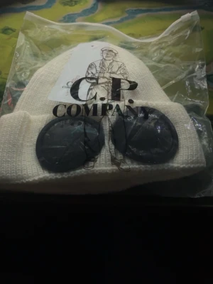 Vit mössa med goggles från C.P. Company - Säljer en vit ribbstickad mössa från C.P. Company med deras ikoniska svarta goggles framtill. Mössan har uppvikt kant och är tillverkad i ett mjukt, stickat material. Perfekt för dig som vill sticka ut med en unik accessoar.