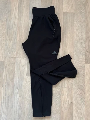 Träningsbyxor från Adidas med dragkedja - Svarta träningsbyxor från Adidas med dragkedjor vid bensluten och fickorna för extra prestanda. Byxorna har en bred resår i midjan och atletisk passform. Diskret Adidas-logga på vänster lår.  Byxorna är tillverkade i ett mjukt och stretchigt material vilket gör dem perfekta för träning (t.ex fotboll, gym) eller bara att ha hemma.  Använda bara fåtal gånger. 