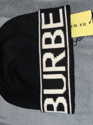 Svart mössa från Burberry med logga - Säljer en svart stickad mössa från Burberry med bred vit kant och stor kontrasterande logga runtom. Klassisk beanie-modell som är både stilren och trendig, perfekt för att lyfta din outfit.